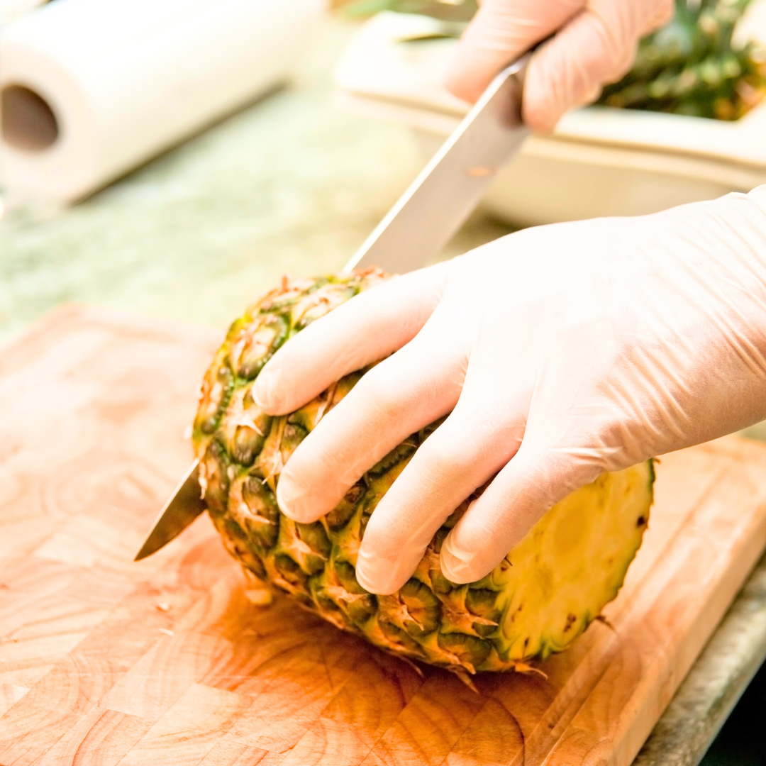 Hände mit Handschuhen schneiden eine frische Ananas auf einem Schneidebrett, saftig, naturbelassen für die Herstellung von LinoraBel Seifen.