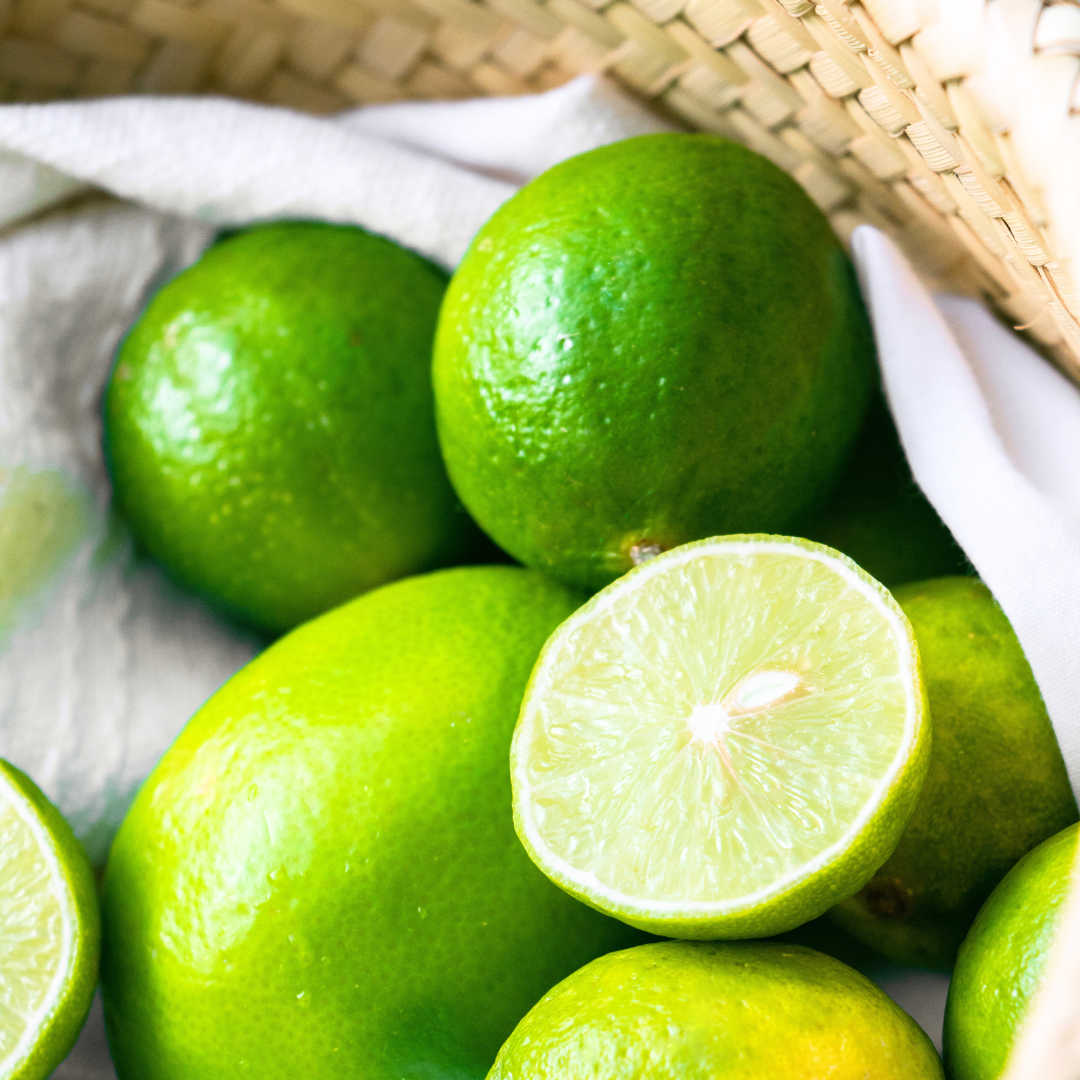 Frische Limetten in einem Korb, darunter eine halbierte Limette mit saftigem Fruchtfleisch auf weißem Tuch.