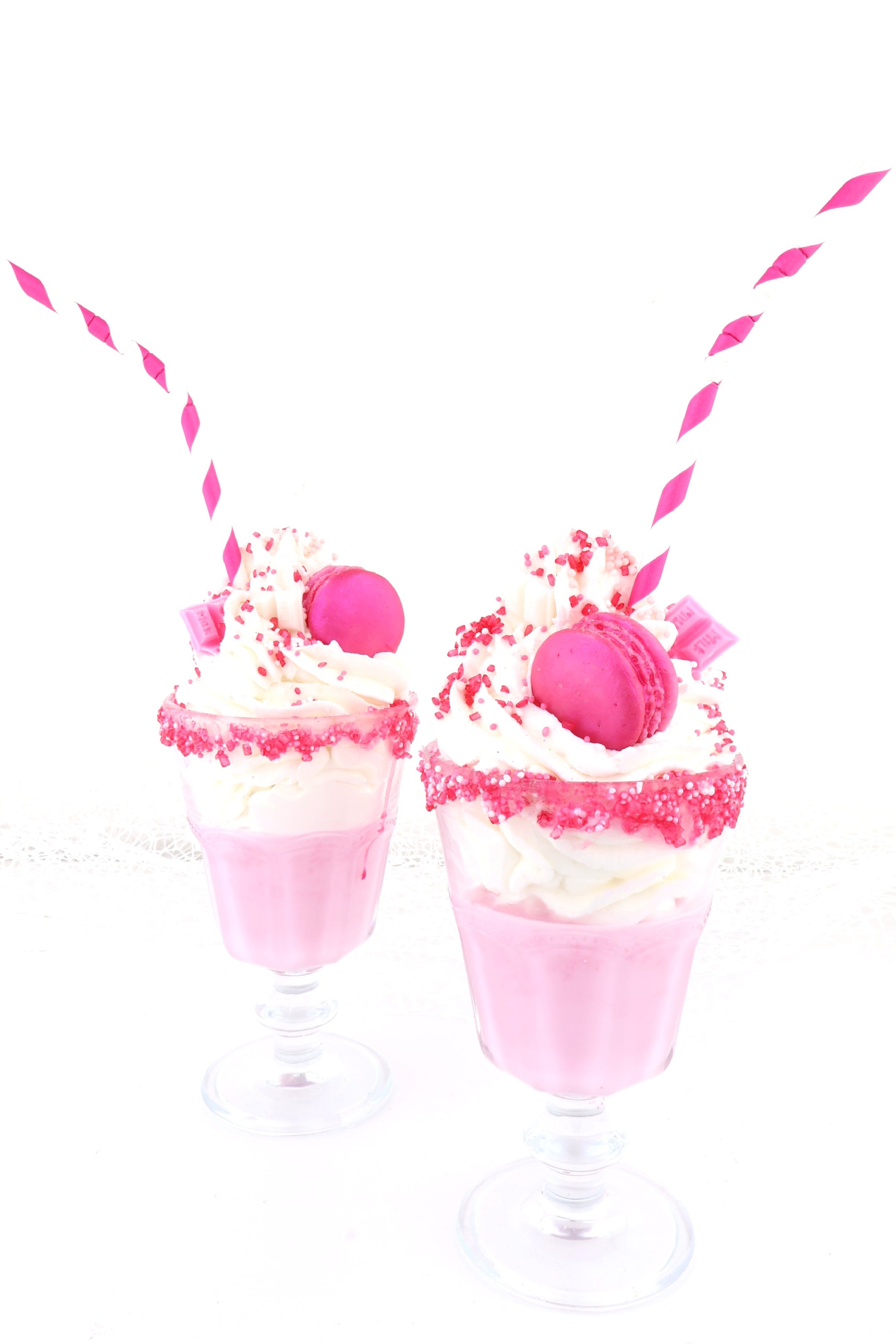 Zwei pink-weiße Milkshake-Dessertkerzen mit Sahnetopping, Macarons, Erdbeeren, Schokostücken, Zuckerrand und gestreiften Strohhalmen.